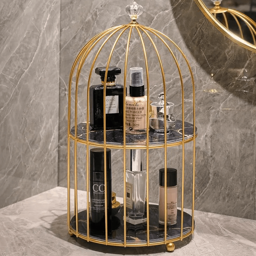 A gold birdcage-style two-tier vanity organizer with black marble trays, holding assorted cosmetic bottles and jars, set against a gray marble wall and counter.