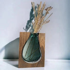 Teal glass vase with dried plants inside, set against a wooden block on a light background