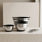 Set of stainless steel mixing bowls with black lids on a neutral background