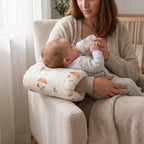 A baby bottle-feeding mother sitting in a cream armchair, supported by a mushroom-patterned nursing pillow.