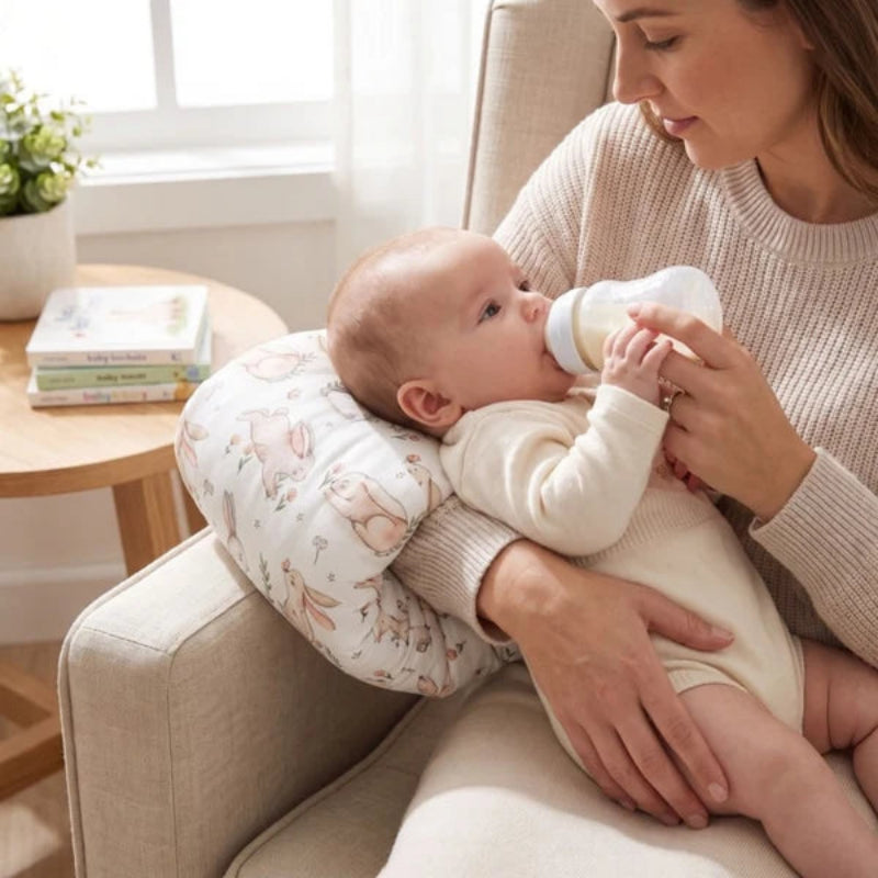A baby wearing a cream bodysuit rests on a woman's lap in a beige armchair, drinking from a white bottle while supported by a patterned baby support pillow.