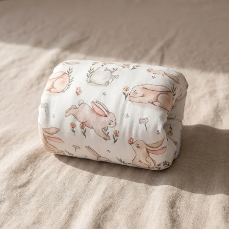 A folded baby blanket with a bunny print, resting on a textured beige linen surface under soft natural sunlight.