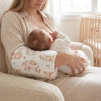 A woman breastfeeding a baby while wearing a white nursing pillow with a pink and beige bunny pattern. The scene is set in a cozy, softly lit room with a wooden crib in the background.
