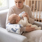 A baby bottle-feeding mother sitting on a light gray armchair, using a whale-print baby support pillow to support her baby.