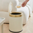 Beige trash bin with gold rim next to a white couch, with a hand holding a plate of food above it.