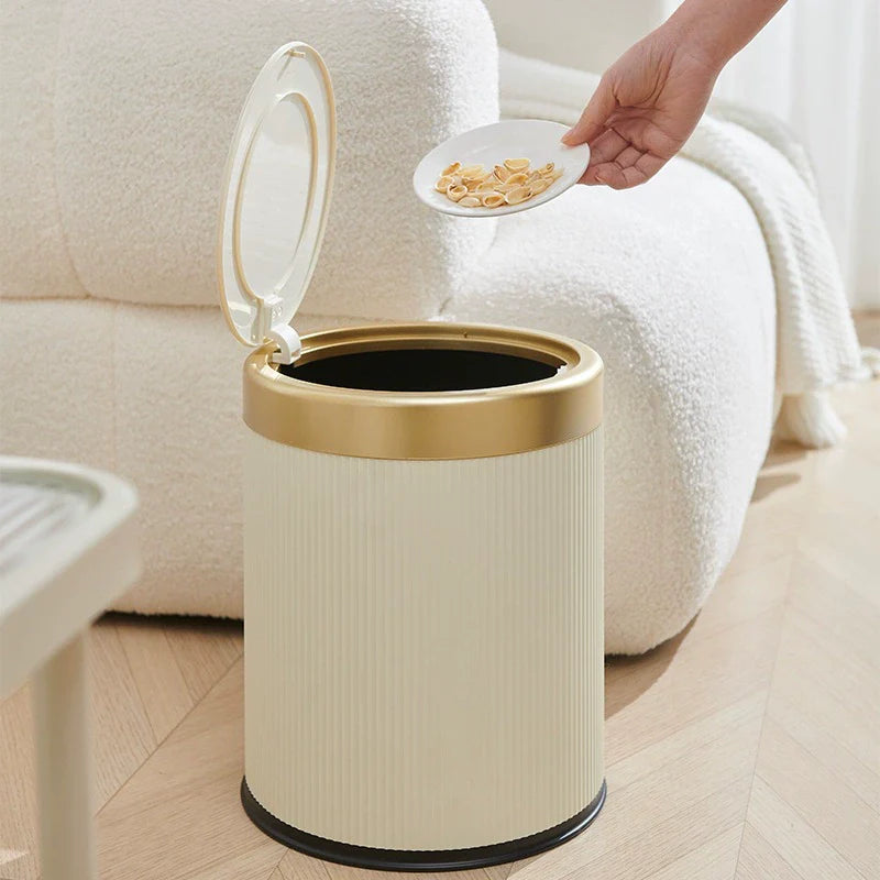 Beige trash bin with gold rim next to a white couch, with a hand holding a plate of food above it.