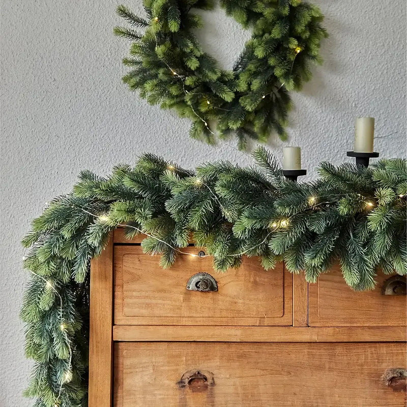 A green pine garland with warm white string lights draped over a wooden chest of drawers, with a matching green wreath hanging on the wall above.