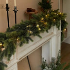 A green garland with warm white string lights draped across a white fireplace mantel in a cozy indoor setting.