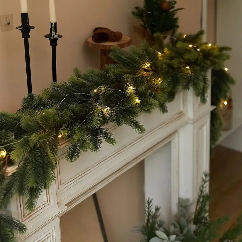 A green garland with warm white string lights draped across a white fireplace mantel in a cozy indoor setting.