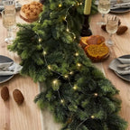 A green pine garland with warm white string lights draped across a wooden dining table, surrounded by plates, wine glasses, bread, and candles in a cozy holiday setting.