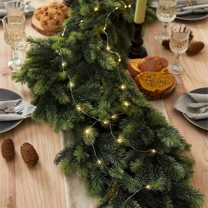 A green pine garland with warm white string lights draped across a wooden dining table, surrounded by plates, wine glasses, bread, and candles in a cozy holiday setting.