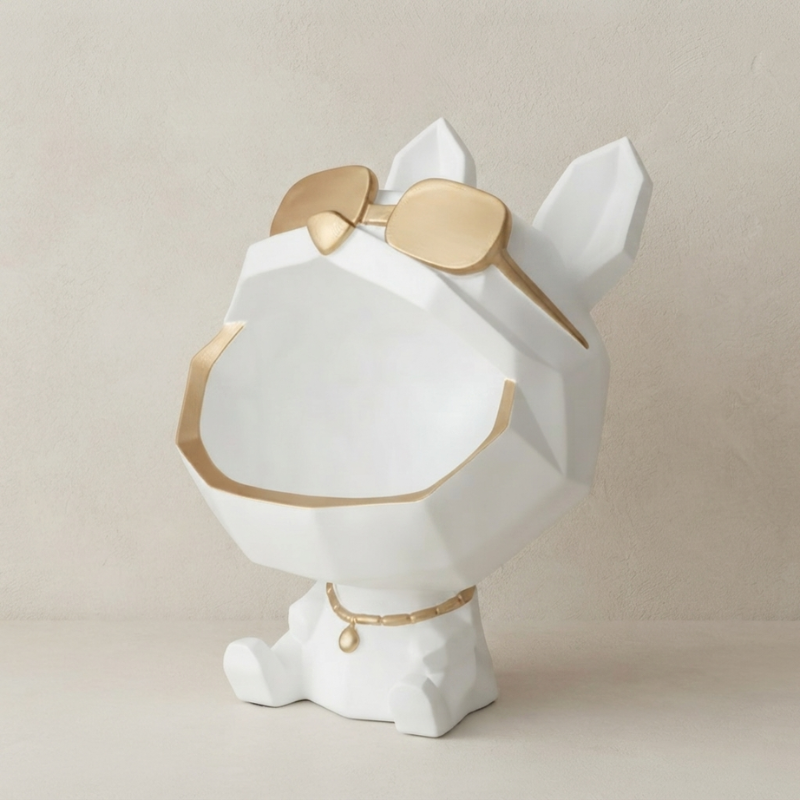 A white geometric dog-shaped decorative object with gold trim, gold sunglasses on its head, and a gold collar with a small gold bead, photographed against a warm beige studio background.