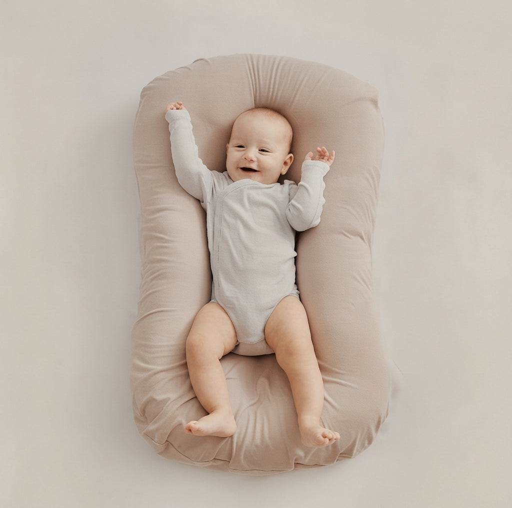A baby lying on a beige padded bassinet or changing pad against a plain light beige background. The baby is wearing a light gray long-sleeve bodysuit and has one arm raised in the air.