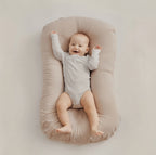 A baby lying on a beige padded bassinet or changing pad against a plain light beige background. The baby is wearing a light gray long-sleeve bodysuit and has one arm raised in the air.