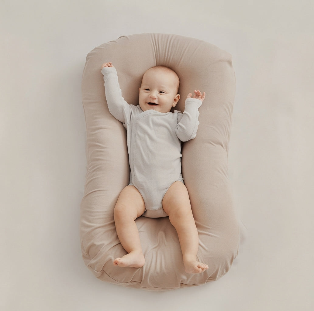A baby lying on a beige padded bassinet or changing pad against a plain light beige background. The baby is wearing a light gray long-sleeve bodysuit and has one arm raised in the air.