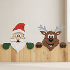 A festive holiday wall decoration set featuring a smiling Santa Claus head with a red hat, white beard, and green mittens on the left, and a brown reindeer head with gray antlers on the right, both peeking over a wooden panel against a light beige wall.