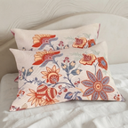Two decorative throw pillows with a colorful floral and bird pattern on a cream background, styled on a white bed.