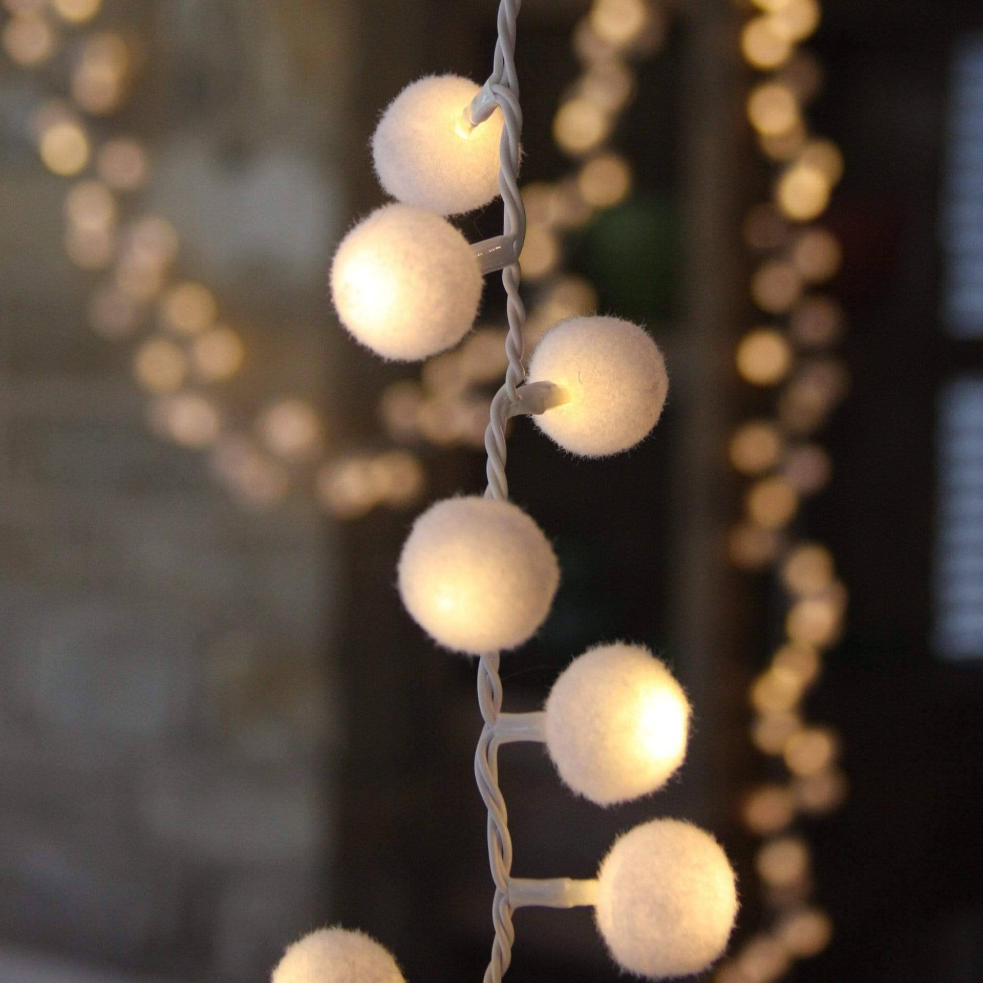 Warm white LED string lights with fluffy pom-pom bulbs hanging vertically against a dark, softly blurred background of more bokeh lights.