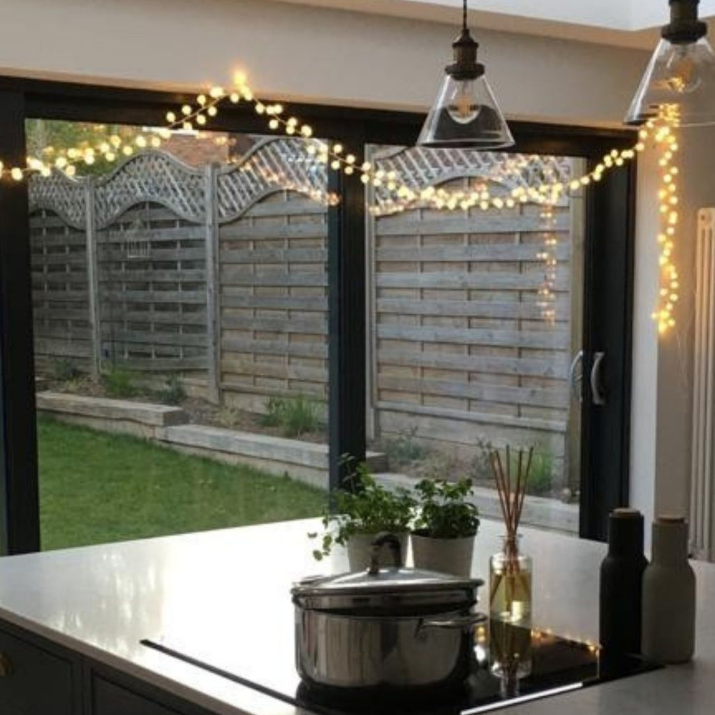 Warm white string lights draped across a black-framed sliding glass door in a modern kitchen, with a wooden fence and green lawn visible outside. A stainless steel pot sits on a black stovetop on the white countertop.