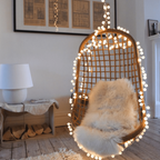 A hanging wicker egg chair with a fluffy cream sheepskin cushion, decorated with warm white string lights, set in a cozy room with a wooden floor and a white side table.