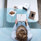 Person sitting at a glass table with a book, croissant, and coffee.