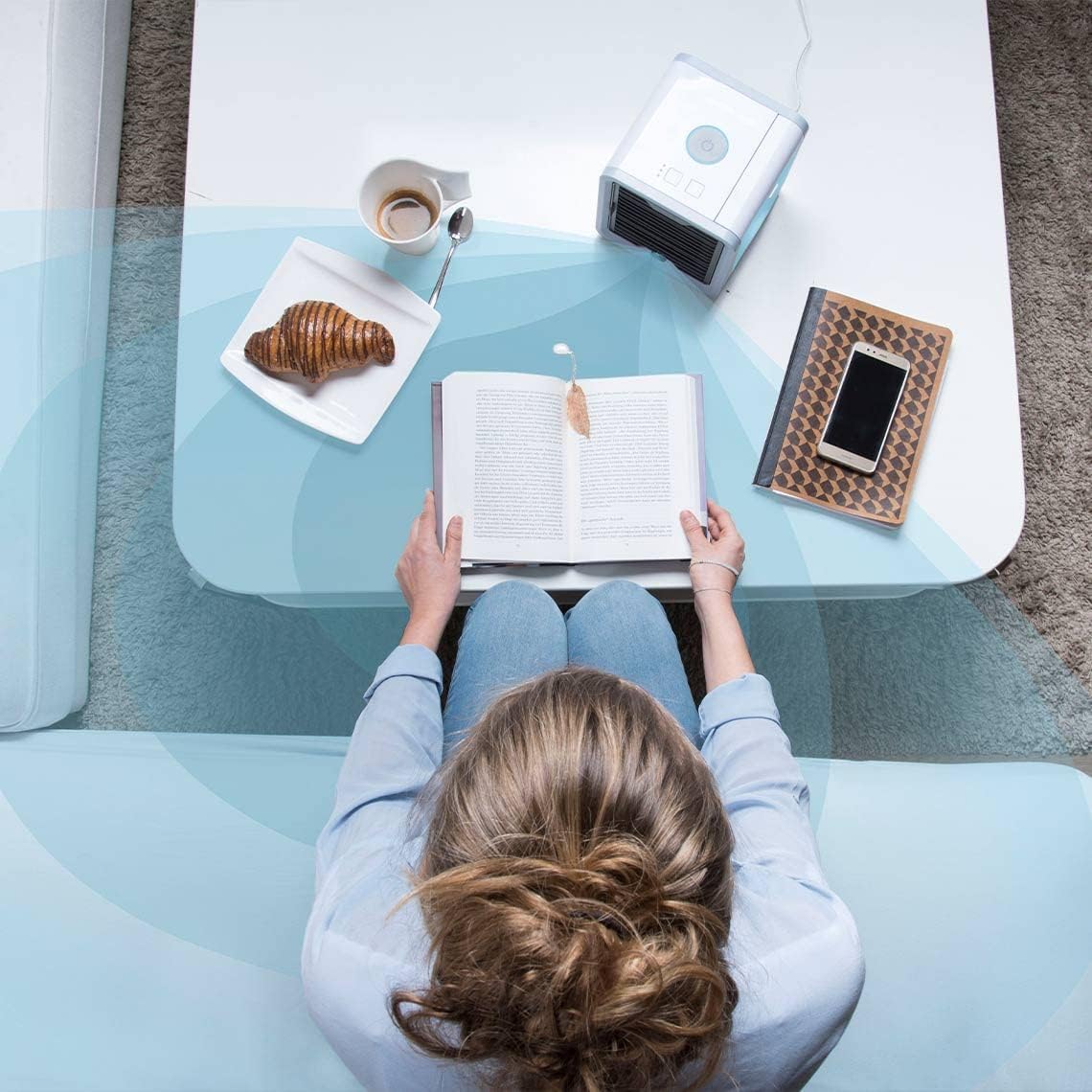 Person sitting at a glass table with a book, croissant, and coffee.