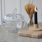 A clear glass pumpkin-shaped container with a ribbed texture and stem, styled on a light wooden table next to a black candle in a wooden holder and dried wheat in a glass vase.