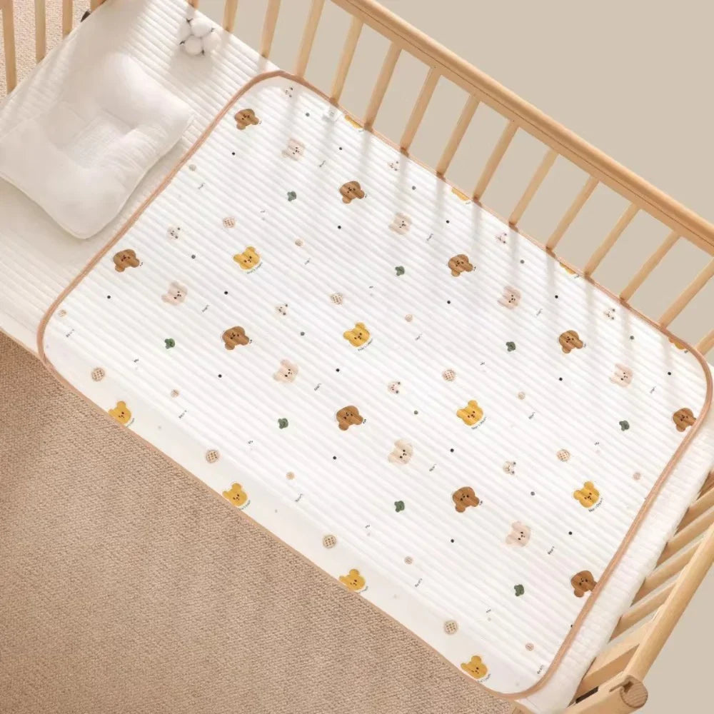 A white baby mattress protector with a cute teddy bear and flower pattern, shown fitted inside a light wood crib with a white pillow.