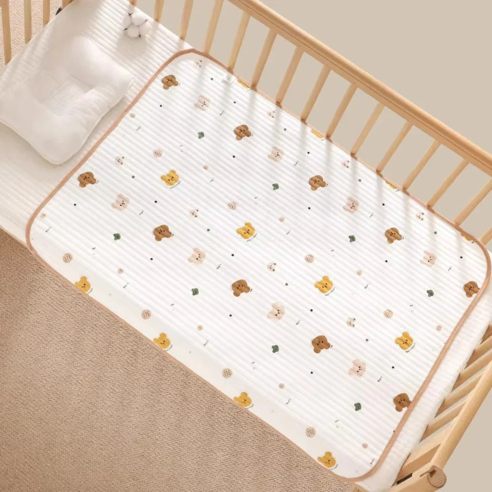 A white baby mattress protector with a cute teddy bear and flower pattern, shown fitted inside a light wood crib with a white pillow.