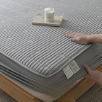 A close-up of a gray quilted mattress topper or mattress cover on a bed frame, with a person's hands touching the fabric and a small candle resting on top. The product has a ribbed quilted texture and a visible care label near the corner.