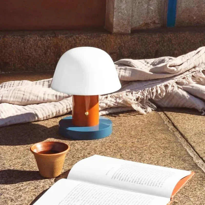 White lamp on a blue base next to an open book and small pot on a concrete surface.