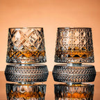 Two crystal glasses filled with a amber liquid on a reflective surface with a peach-colored background.