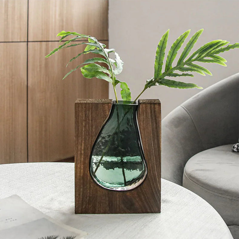 Green plant in a teardrop-shaped glass vase with wooden base on a white surface.