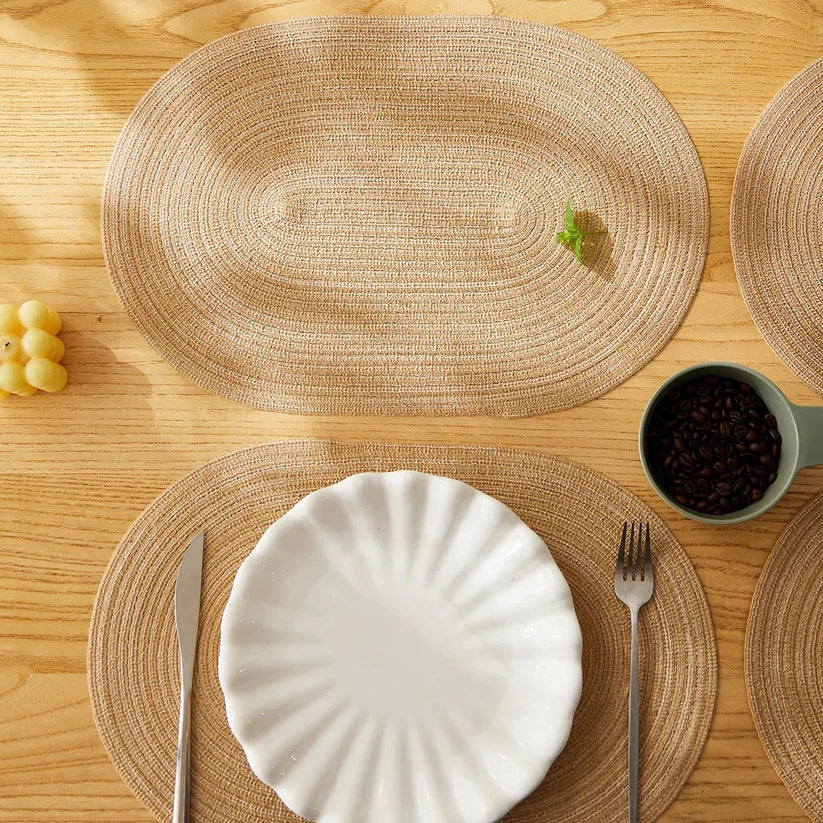 Table setting with woven placemats, white plate, silverware, and a cup on a wooden table.
