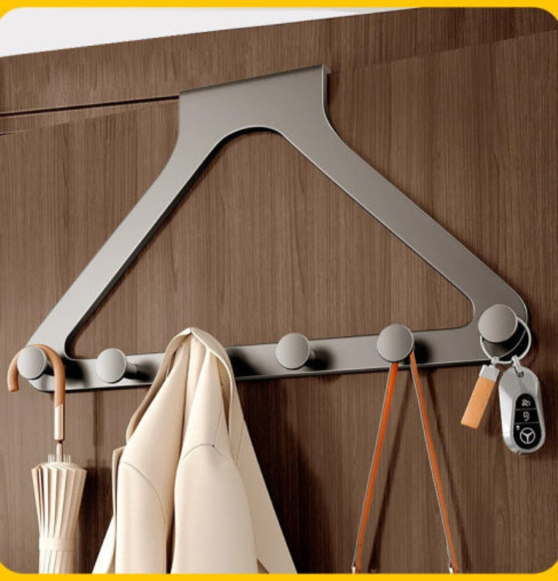 A modern wall-mounted triangular coat rack with five round hooks, shown attached to a brown wooden door. A cream coat hangs from one hook, and keys and straps are attached to the right side.