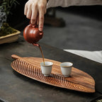 Person pouring tea from a red teapot into two white cups on a wooden leaf-shaped tray.