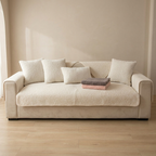 Beige sofa with cushions and a blanket on a wooden floor against a beige wall.