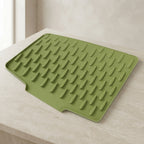 An olive green silicone dish drying mat with raised drip edges, shown resting on a light beige stone countertop against a matching tiled wall.