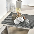 Gray kitchen mat on a countertop with various items including a white cup, whisk, and glass.