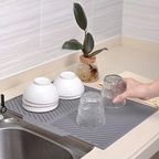 Gray dish drying mat on a kitchen counter with dishes and a plant in the background.