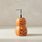 An orange Halloween-themed soap dispenser shaped like two stacked jack-o'-lantern pumpkins with a shiny silver metal pump, set against a neutral beige background.