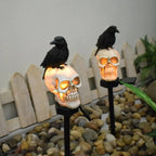 Two skull-shaped lights with crows on top, placed on a gravel surface with a wooden fence in the background.
