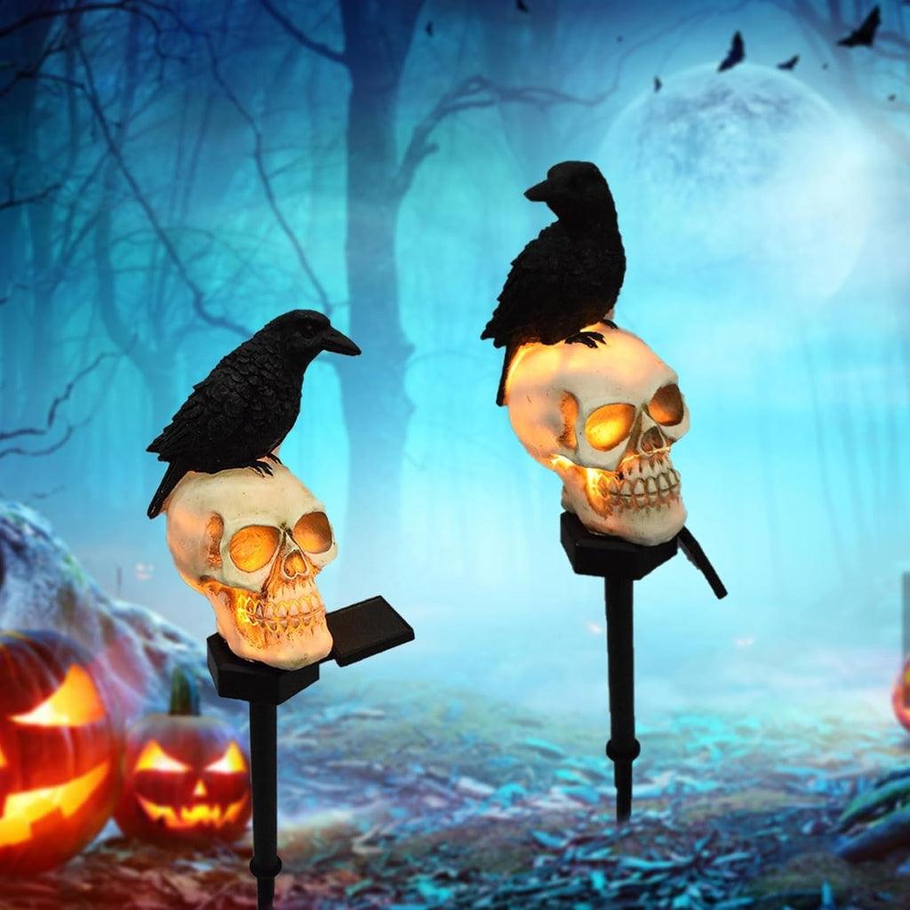 Two Halloween yard lights shaped like skulls with black crows perched on top, shown against a spooky blue forest background with jack-o'-lanterns and a full moon.