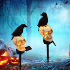 Two Halloween yard lights shaped like skulls with black crows perched on top, shown against a spooky blue forest background with jack-o'-lanterns and a full moon.