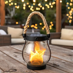 Solar lantern with flame effect on a wooden table outdoors, surrounded by blurred lights and furniture.