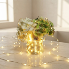 Warm white LED string lights wrapped around a vase filled with white and green hydrangea flowers, set on a light-colored surface in a softly lit room.