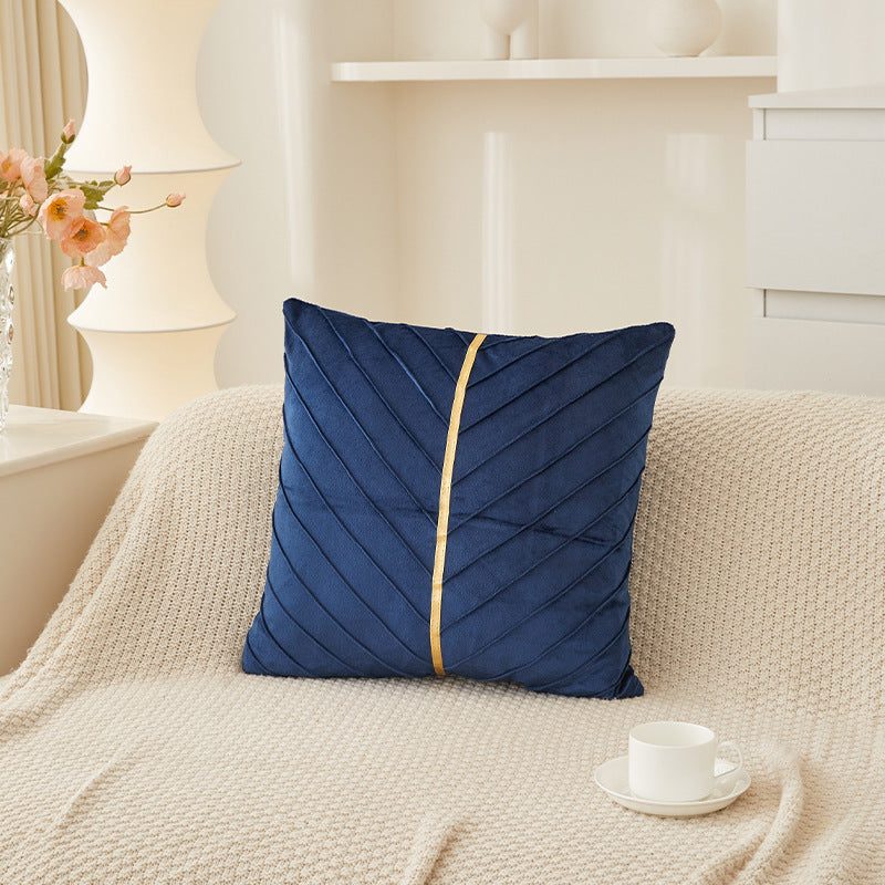 A navy blue square throw pillow with a vertical gold stripe and diagonal pleated texture, resting on a cream-colored knit sofa in a bright, modern living room setting.