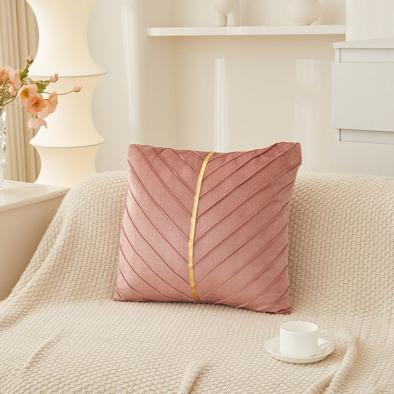 A square pink throw pillow with a gold center stripe and pleated chevron texture, styled on a cream knit blanket in a softly lit living room setting.
