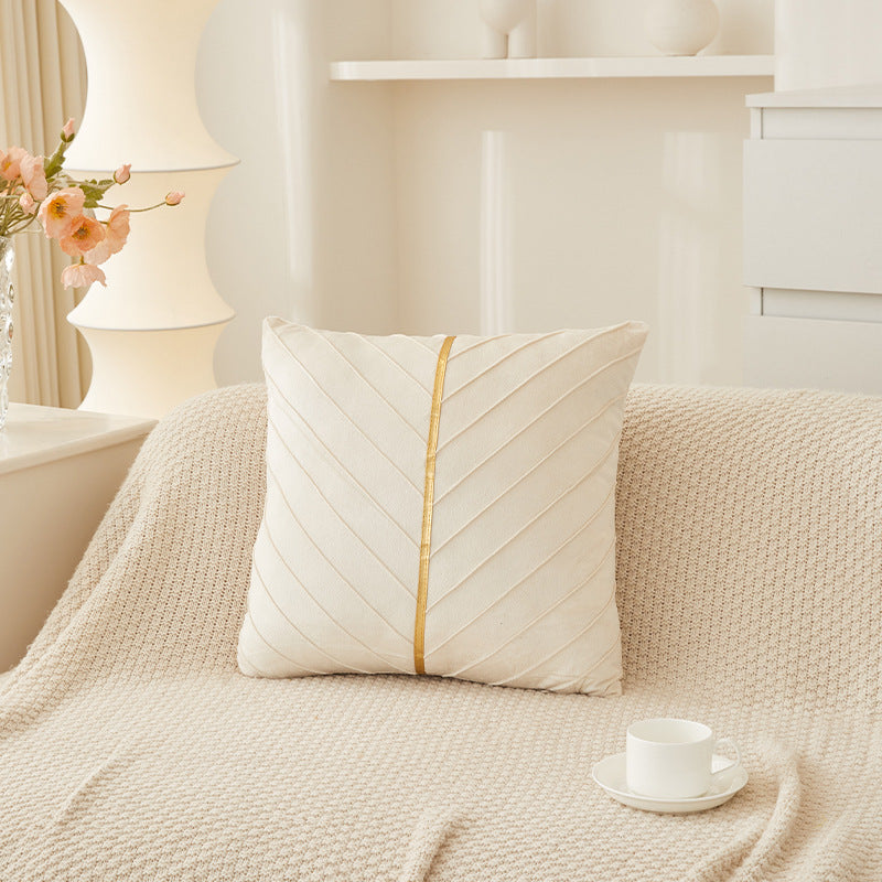A square cream throw pillow with a central gold stripe and diagonal stitched texture, resting on a textured beige knit blanket in a softly lit, minimalist room.
