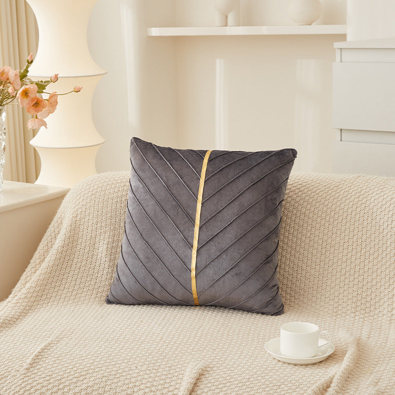 A square gray throw pillow with a gold vertical stripe and stitched leaf pattern, resting on a cream knit blanket in a softly lit living room setting.